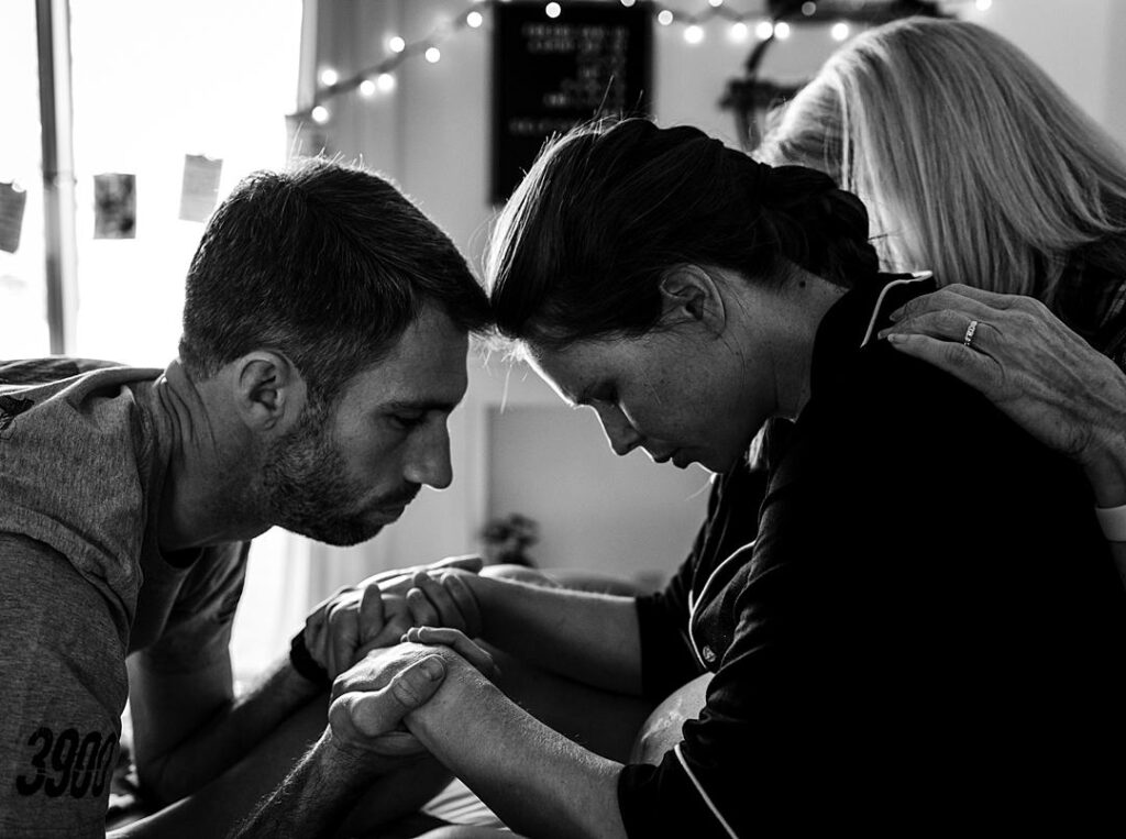 A husband is holding his wife's hands as she leans forward against him through a contraction while her mother sits behind her encouraging her. 