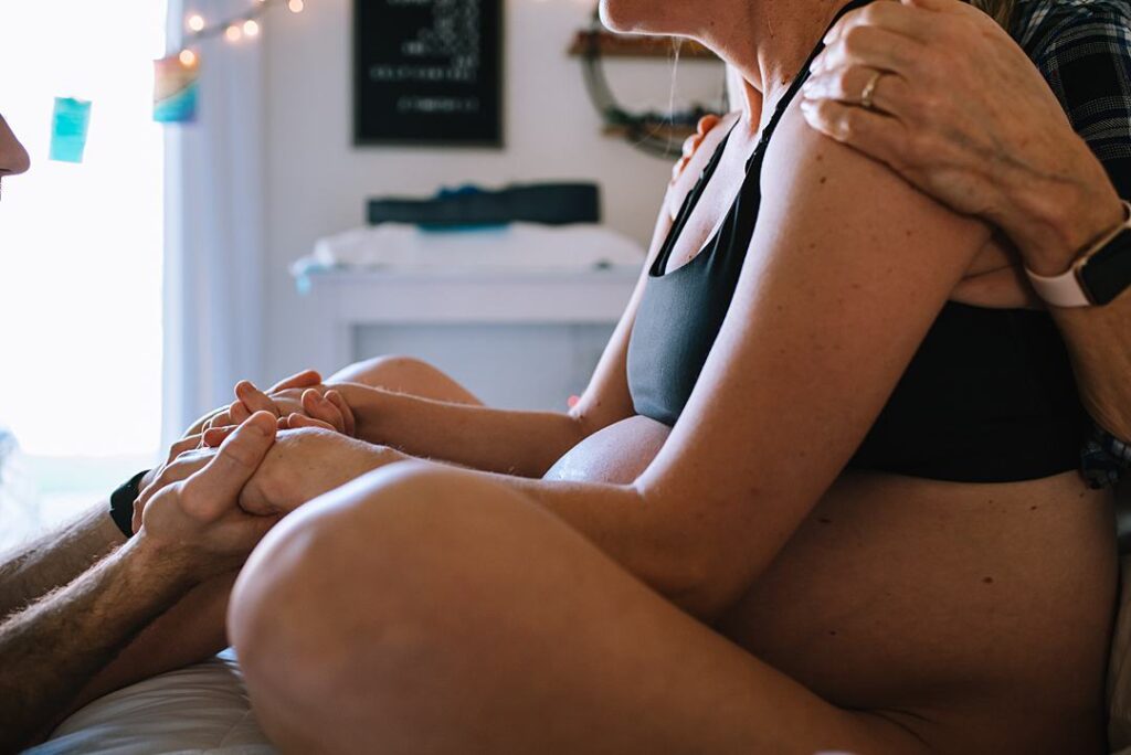 A woman is sitting on her bed at home in labor holding her husbands hand in front of her and her pregnant belly is visible.