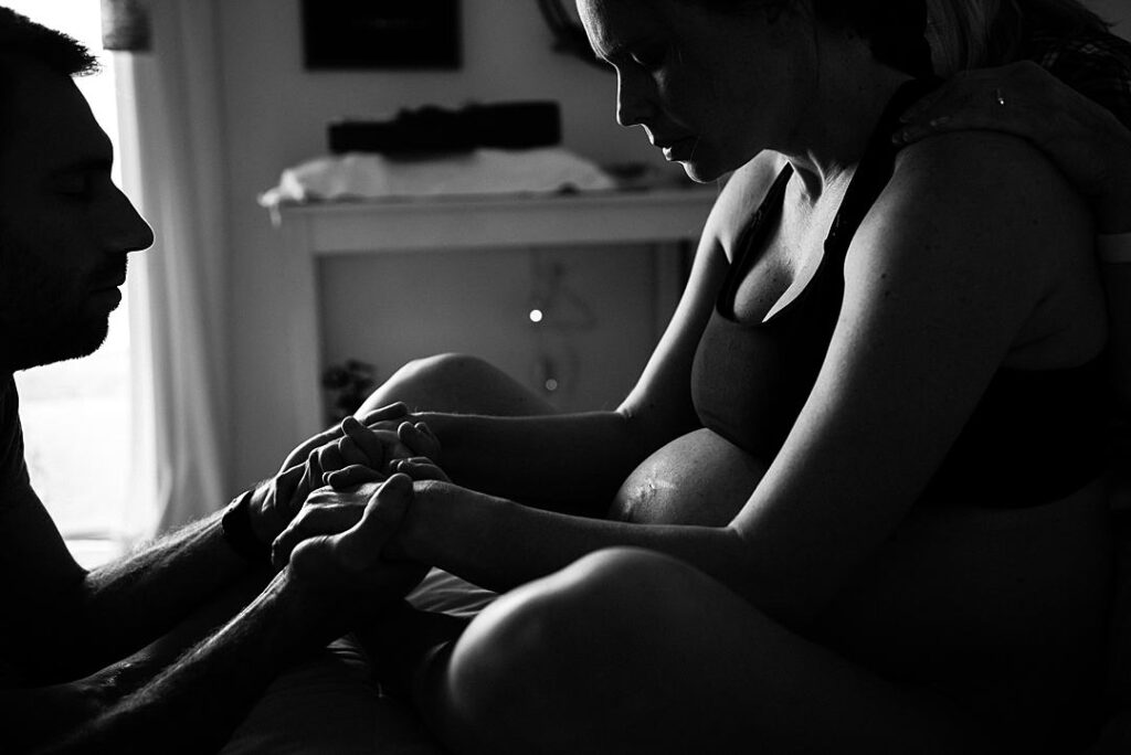 A woman is sitting on her bed at home in labor holding her husbands hand in front of her and her pregnant belly is visible.