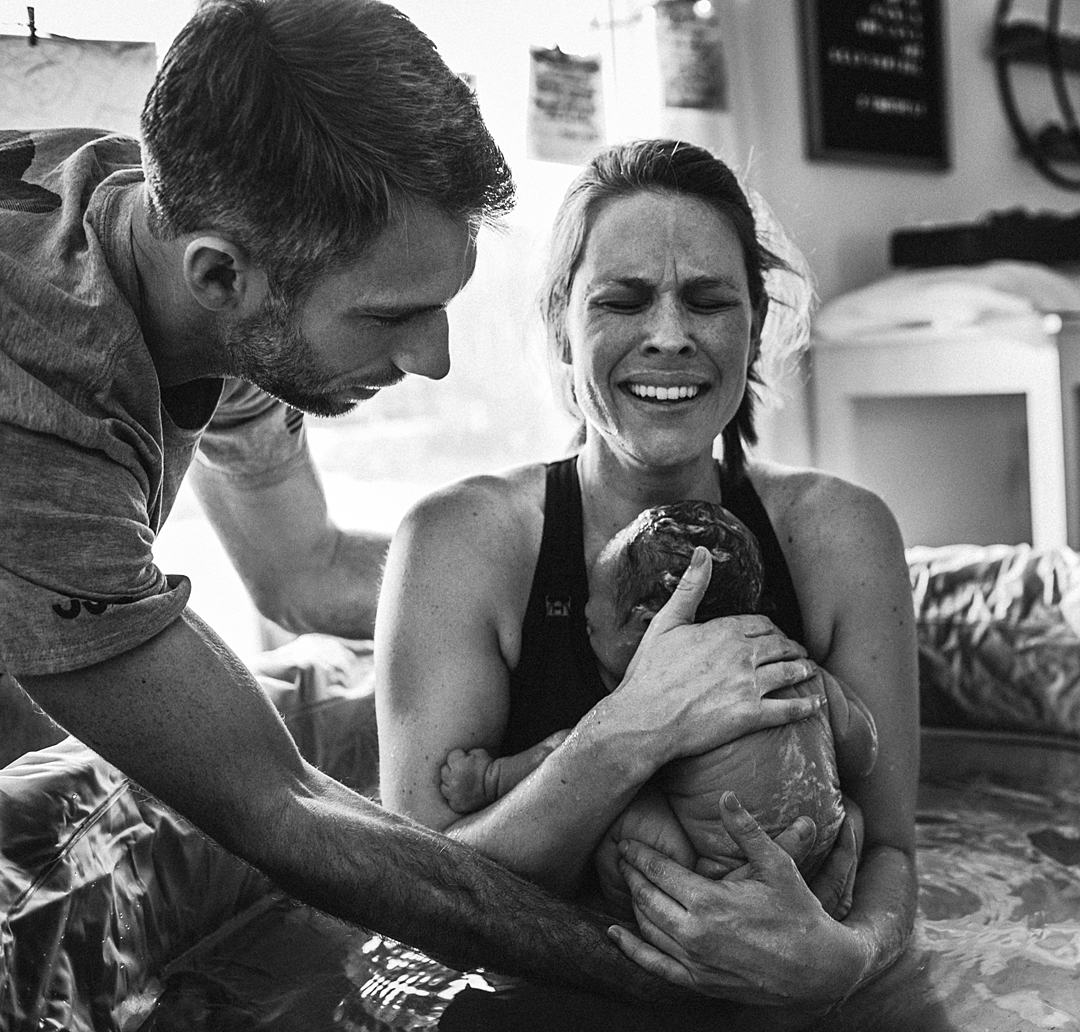 Mother is overjoyed with her just born baby on her chest still sitting in the birth pool at her home birth. Her husband helps to hold the newborn baby with her.